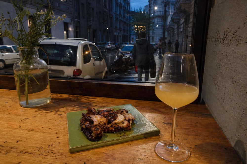 A ceramic-tiled small-plate dish on a wooden counter with a glass of natural wine beside it