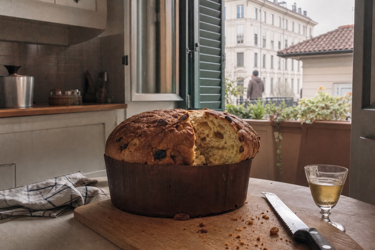 A tall domed panettone on a wooden board in a Milan kitchen, a long serrated knife and a small glass of sweet moscato beside it