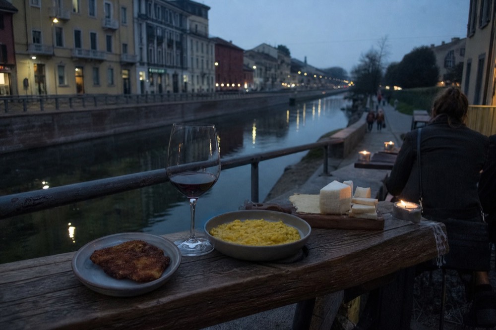 A tasting table with a small breaded cutlet, risotto, cheese, and a glass of red wine