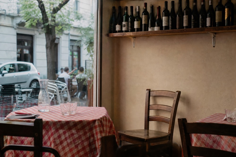 A trattoria dining room with checkered tablecloths, simple wooden chairs and shelves of wine bottles