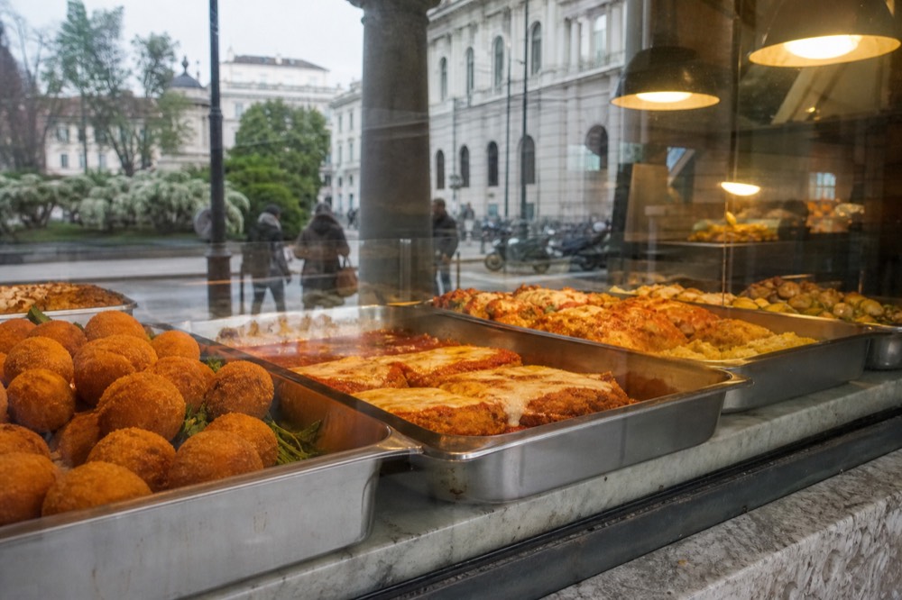 A marble counter with trays of prepared Milanese dishes behind a market glass screen