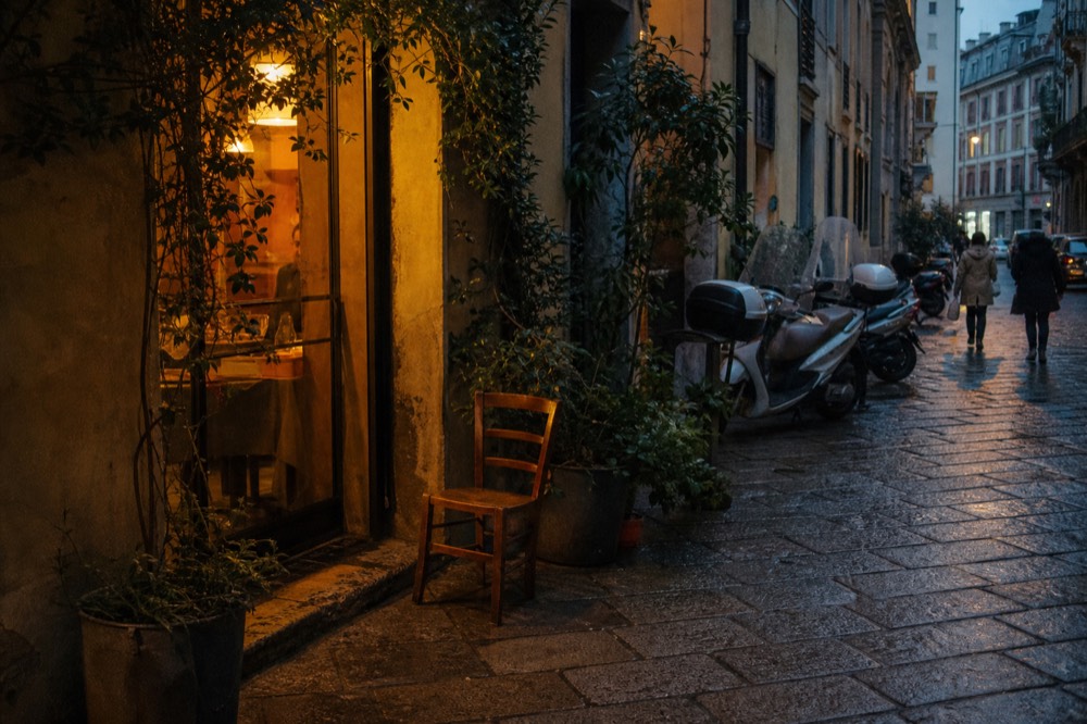 A narrow Milan side street at dusk in October, a small restaurant doorway glowing amber over wet cobbles