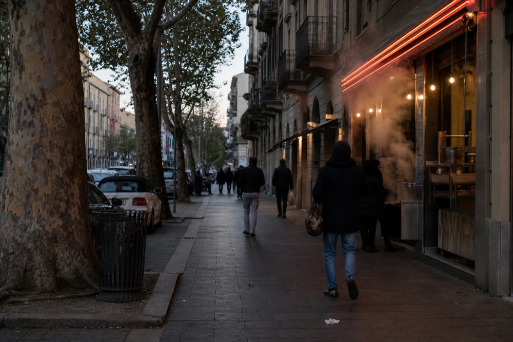 A busy pedestrianised Milan street in the early evening with small food shopfronts and neon signs