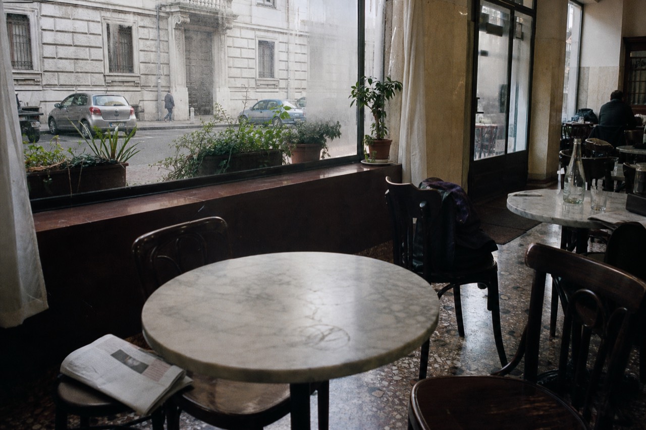A small neighbourhood trattoria interior with marble tables, wooden chairs and a chalkboard menu in Milan