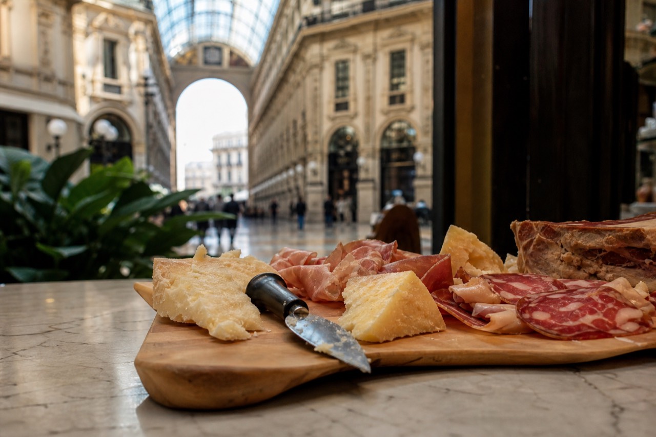 A board of cheeses and cured meats on a marble deli counter, with the glass and iron vault of Milan's Galleria rising behind it