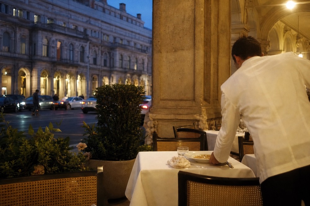 A waiter's hands clearing a plate with leftover pasta from a restaurant table