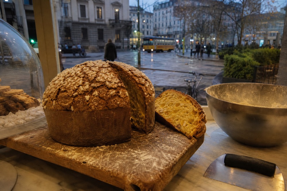 A tall domed panettone with a cracked top crust on a wooden board in a pastry shop