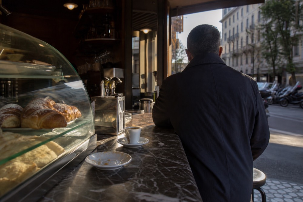 A Milanese bar interior in early morning light, with pastries under a glass case and a customer standing at the counter