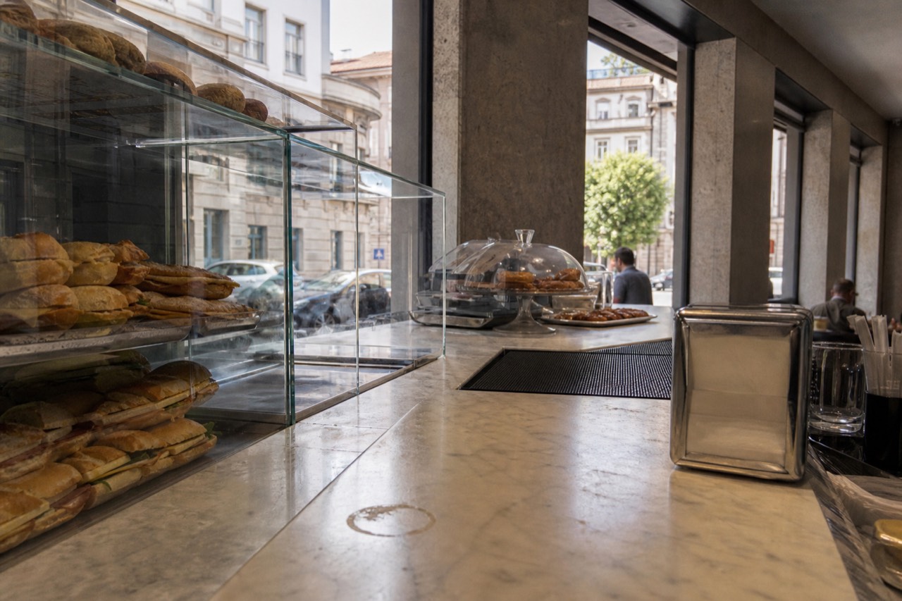 A marble café counter in Milan with glass cases of panini and trays of pastries at midday