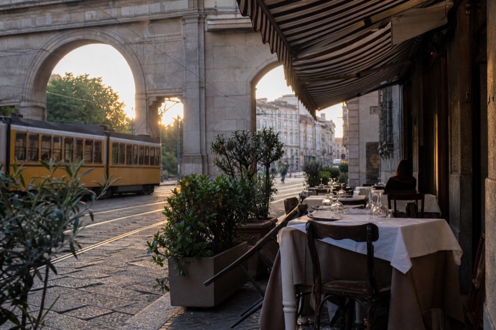 A quiet Milan street in early evening with restaurant tables set under an awning