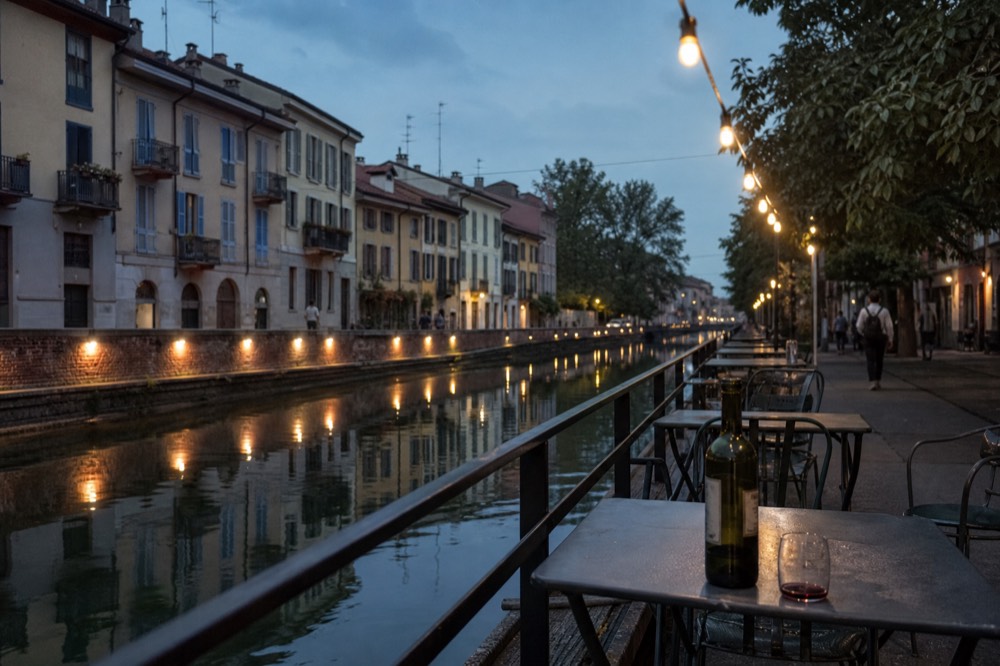 A Navigli canal terrace in the evening with string lights, empty chairs and small tables along the water