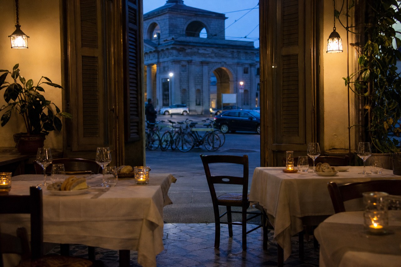 A softly lit Milanese restaurant dining room with empty tables set for dinner, wine glasses and bread baskets.