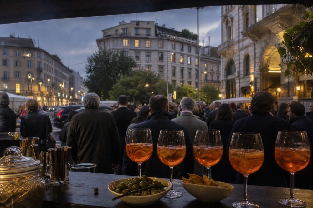 A crowded bar counter with orange spritz glasses and bowls of snacks at dusk