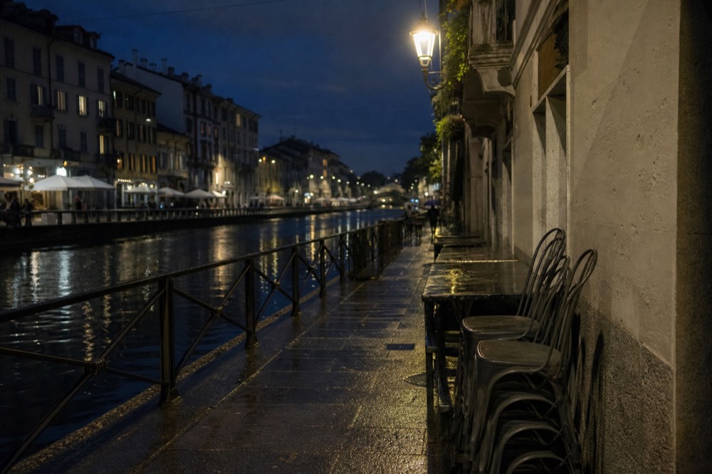 Empty outdoor cafe tables along a Milan canal at night with lit windows behind