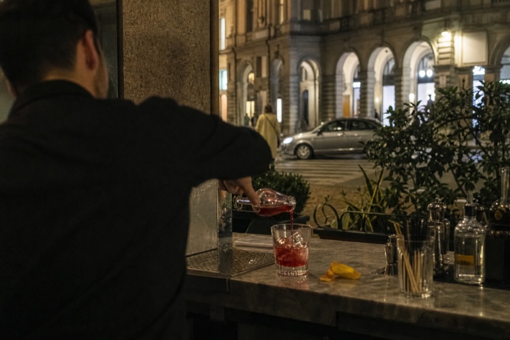 A barman pouring a red bitter cocktail over ice at a dimly lit counter