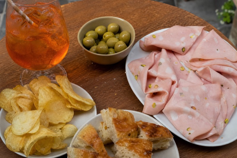 Two spritz glasses with olives, focaccia and mortadella on a canal-side table in Navigli at dusk