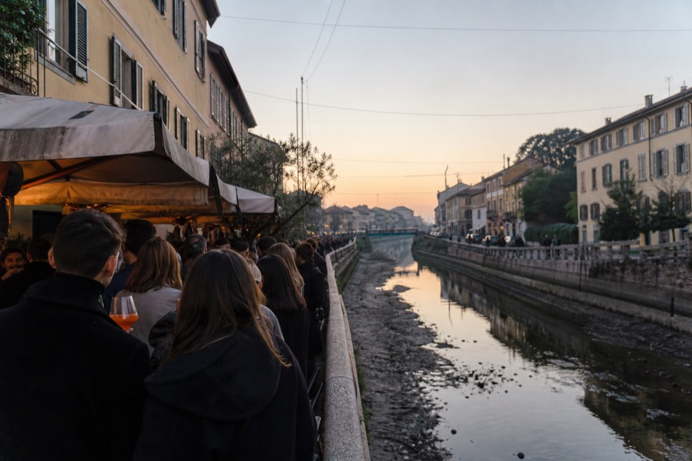 Crowd standing with drinks at an outdoor aperitivo bar along a Milan canal at dusk