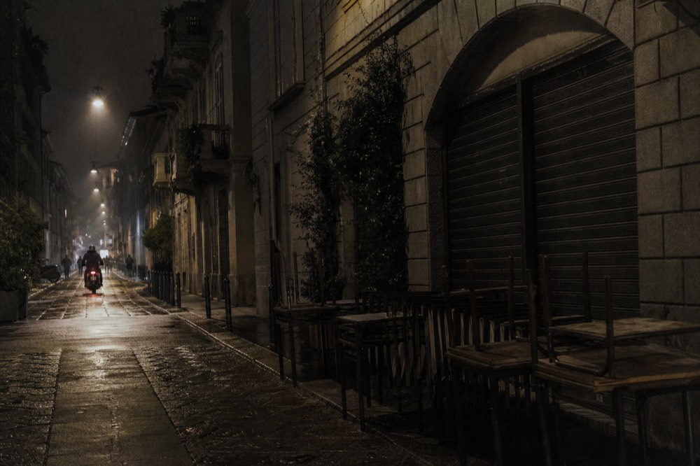 Evening street scene with closed bar shutters and empty outdoor tables in a Milan district