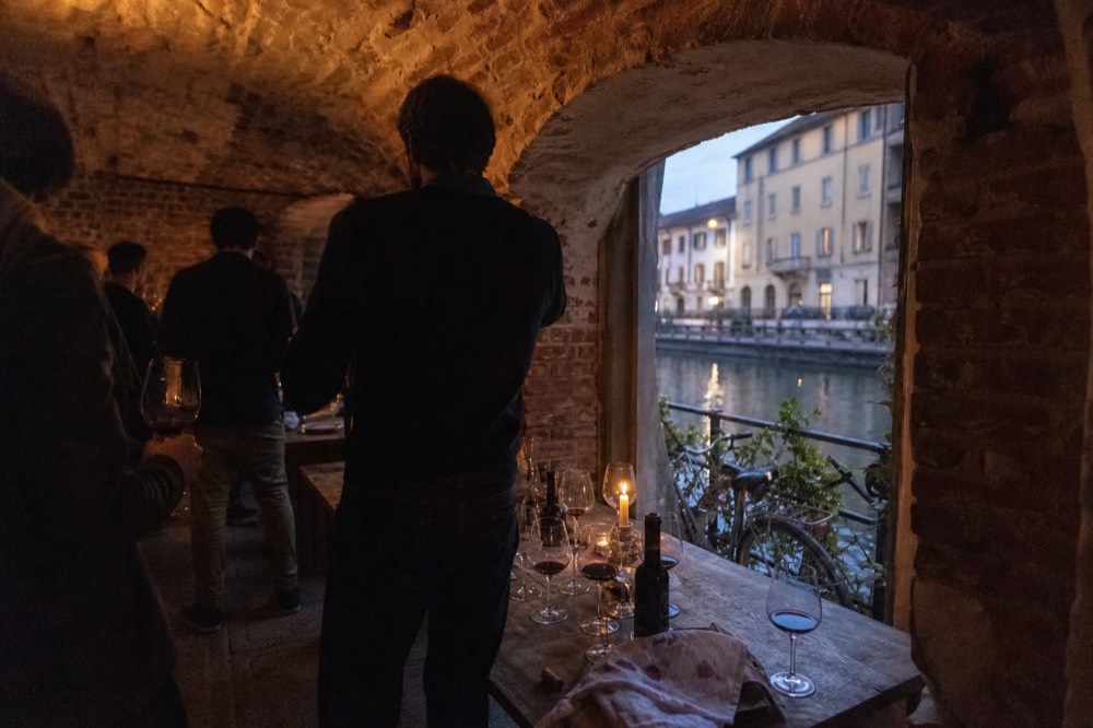 Group of adults with wine glasses standing around a tasting table in a brick-vaulted cellar