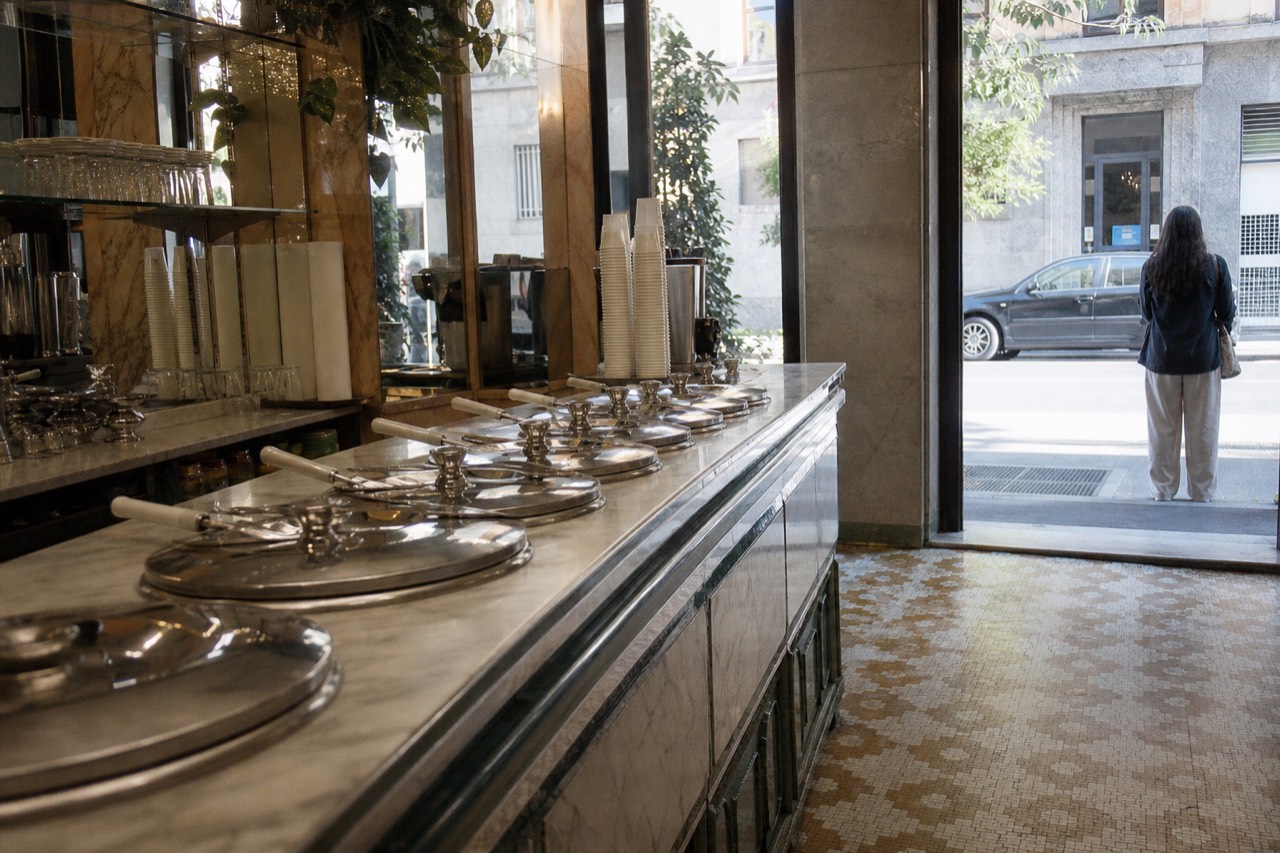 Row of covered metal gelato containers on a marble counter in a small Milanese shop