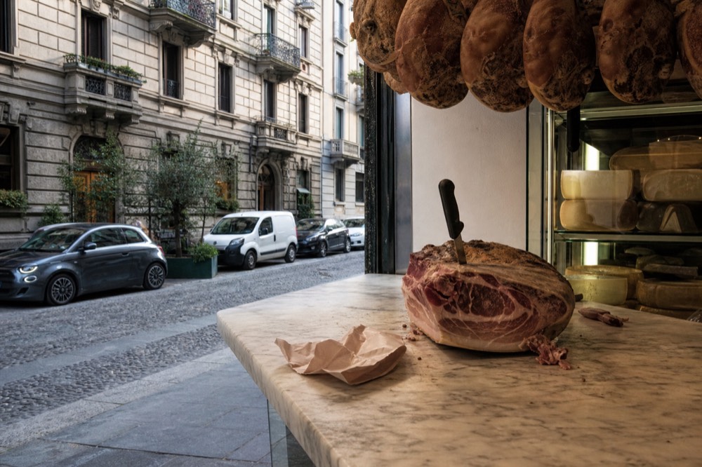 Cured hams hanging above a marble delicatessen counter with cheese wheels stacked behind glass