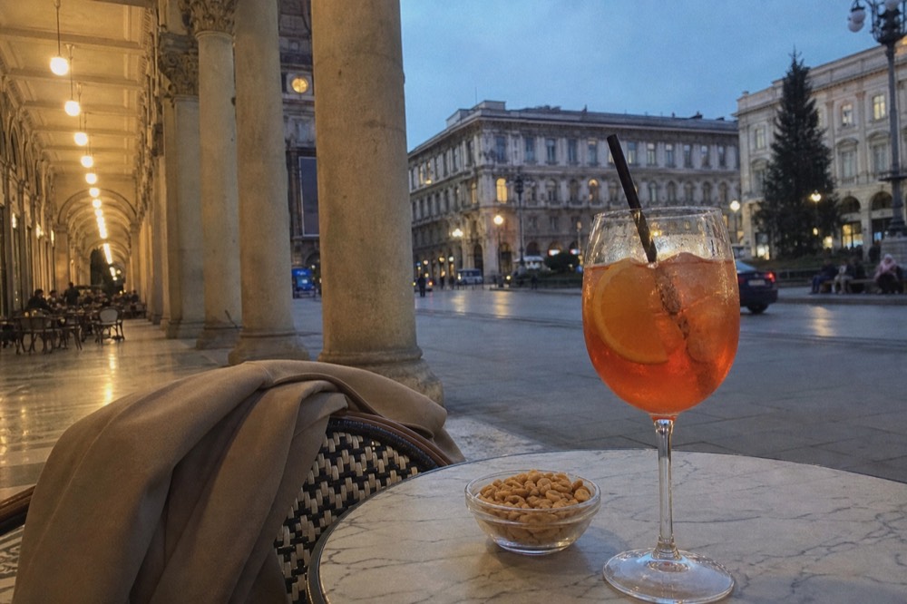 An aperitivo table with a spritz and small snacks beside a Milan arcade at dusk