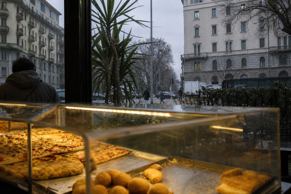Glass display case with rectangular pizza slices and fried snacks in a Milan bakery