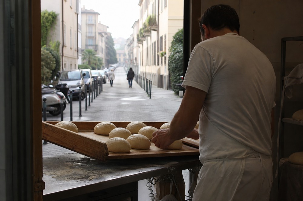 Baker placing bread rolls on a wooden tray inside a Milan bakery