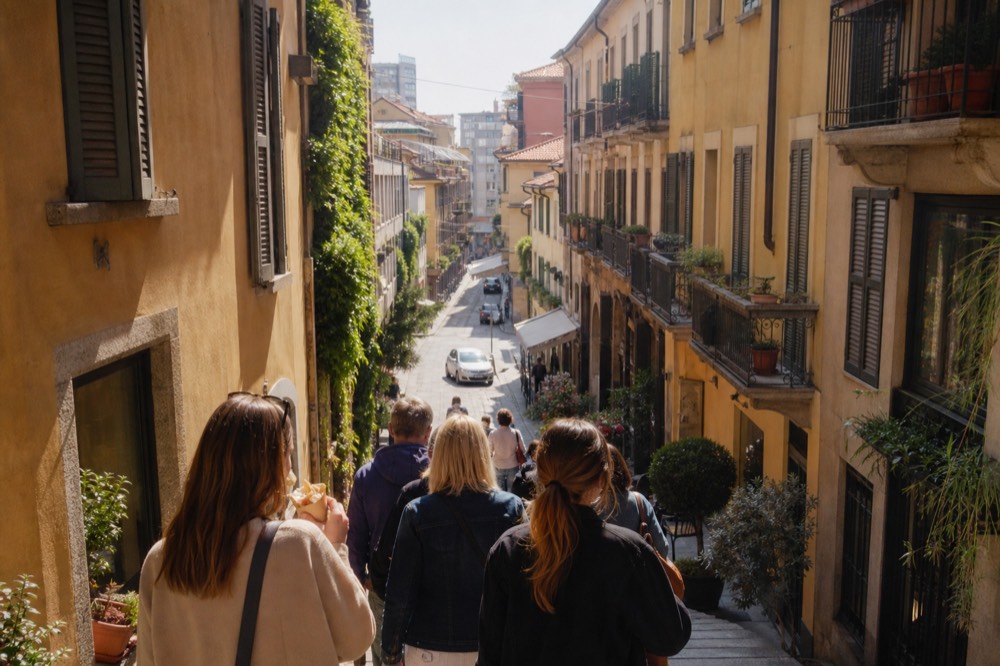 Small group of visitors following a guide along a Milan street with food shops