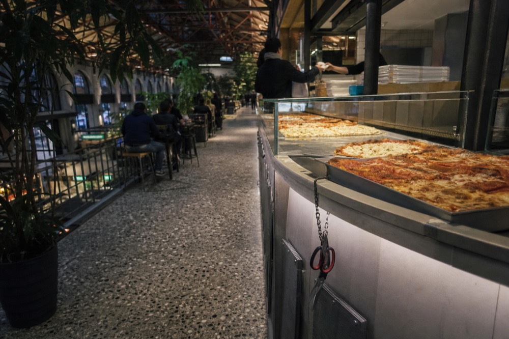 A market food hall counter with trays of rectangular pizza and a price board