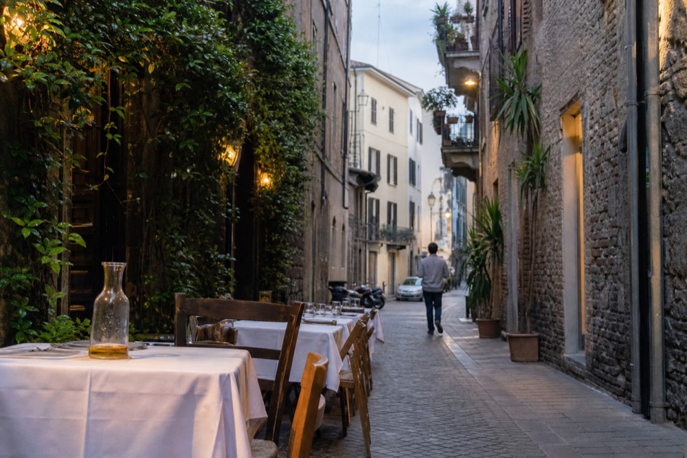 A narrow Milan street with restaurant tables and chairs set out under building facades