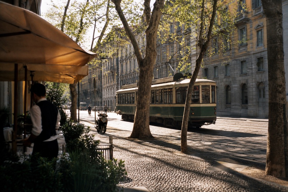A tram passing a Milan street corner with cafe awnings in late afternoon light